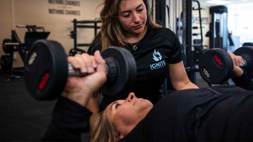 Ignite trainer guiding a woman through a dumbbell chest press during a strength-training session.