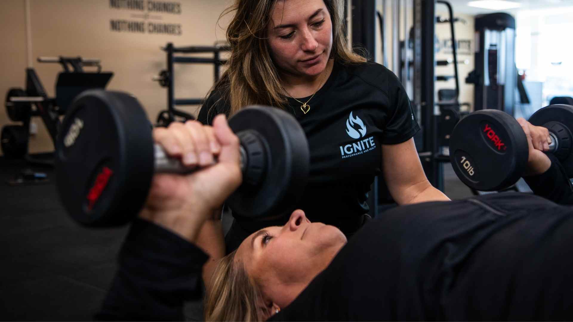 Ignite trainer guiding a woman through a dumbbell chest press during a strength-training session.