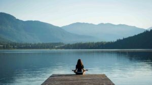 Woman meditating on a dock beside a calm mountain lake.