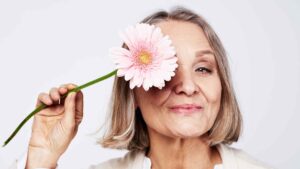 Smiling older woman holding a pink flower over one eye against a light background.