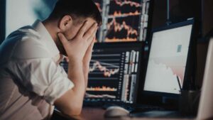 Stressed man sitting at a desk with his face in his hands while looking at computer screens.