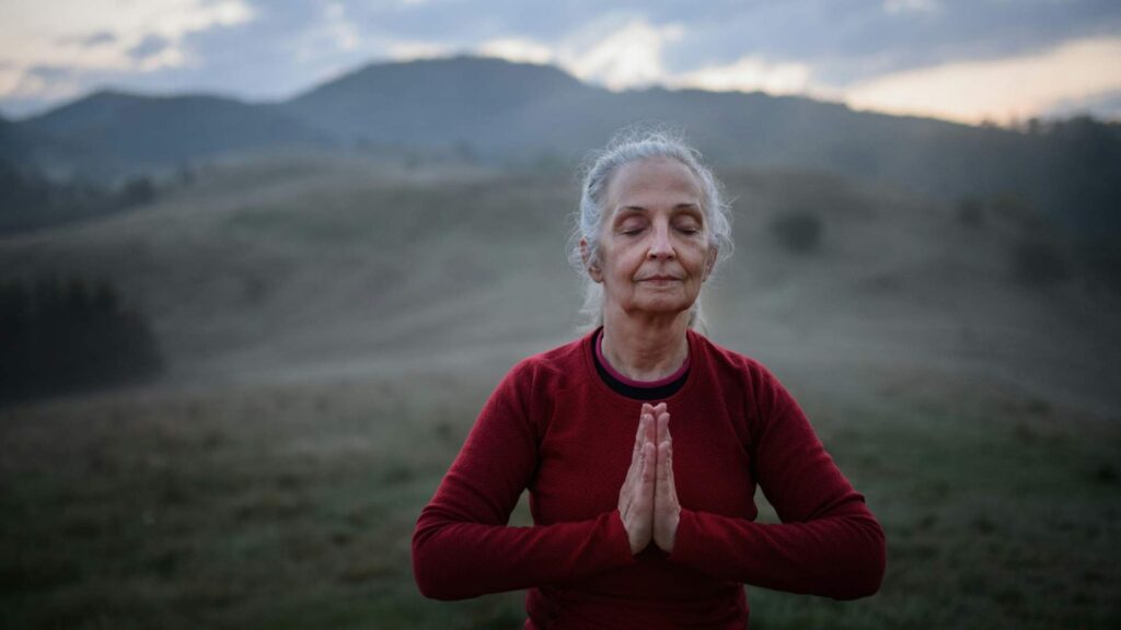 Older woman practicing mindful breathing outdoors with mountains in the background.